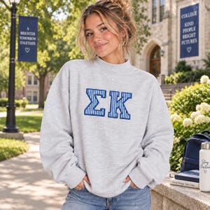 Peut inclure: Un sweat-shirt gris clair à manches longues avec un col montant. Le centre de la poitrine présente les lettres grecques Sigma et Kappa brodées en bleu vif avec un motif à fines rayures verticales blanches sur l'ensemble des lettres.