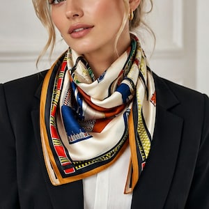 May include: A silk scarf with a cream background and a colorful design featuring buildings and architectural elements. The scarf has a border of black, orange, and gold. The scarf is worn over a black blazer.