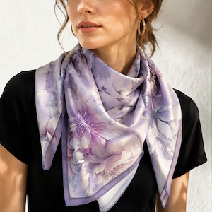 Puede incluir: Un pañuelo cuadrado de seda con un diseño floral en tonos lavanda, morado y blanco. El pañuelo se lleva alrededor del cuello. La persona lleva una camiseta negra. El pañuelo tiene un borde.