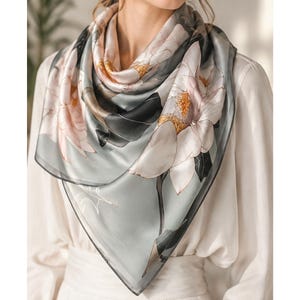 May include: A square silk scarf featuring a floral design with large white and pink flowers on a gray background. The scarf has a black border and is draped around the neck. The scarf is a fashion accessory.