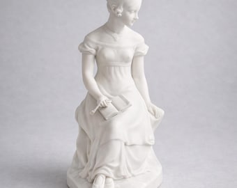 May include: A white porcelain figurine of a seated woman in a long dress, holding a book and a pen. The woman has her hair up and is wearing a necklace. The figurine is on a heart-shaped base.