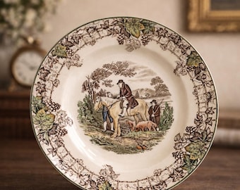 May include: A decorative ceramic plate with a pastoral scene in the center, featuring figures on horseback and dogs. The rim is adorned with a grape and vine pattern in shades of brown and green. The plate has a green border.