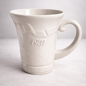 May include: Cream-colored ceramic mug with a curved handle and embossed floral design. The year "1987" is embossed on the side. The mug has a wide rim and a slightly flared shape, suitable for coffee or tea.