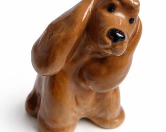 May include: A ceramic figurine of a dog in a sitting position. The dog has long, floppy ears and a black nose. The figurine is a glossy brown color and appears to be a Cocker Spaniel or similar breed.