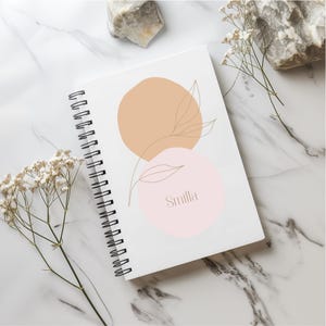Könnte beinhalten: Weißes Spiralnotizbuch mit minimalistischem Design. Das Cover zeigt einen pfirsichfarbenen Kreis, einen rosa Kreis mit dem Namen "Smilla" und filigrane Strichzeichnungen von Blättern. Das Notizbuch liegt auf einer Marmoroberfläche.