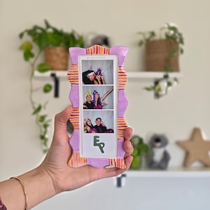 May include: A rectangular ceramic photo frame with a wavy, scalloped edge painted in lavender and orange stripes. The frame holds a vertical photo strip featuring three panels of two people wearing party props and a green logo at the bottom.