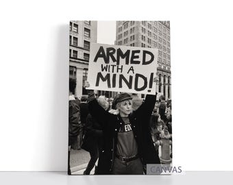 May include: Black and white photograph of a person holding a sign that reads "ARMED WITH A MIND!" in a crowd. The person is wearing a black jacket and a hat. The background includes buildings and other people.
