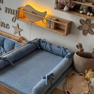Könnte beinhalten: Eine Wickelkommode mit einer hellblauen Wickelauflage und Stoßfängern. Ein Holzregal mit einem vogelartigen Licht und den Buchstaben "DA BY" ist darüber angebracht. Ein geflochtener Korb und ein Häkelstrampler befinden sich an der Seite.