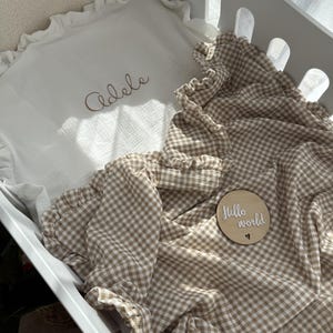 Peut inclure: Un berceau blanc avec un oreiller blanc à volants brodé du nom "Adele" en fil marron. Une couverture à carreaux beige et blanche avec des bords à volants et un disque en bois avec "Hello world".