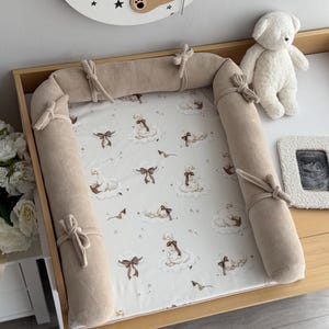 May include: A rectangular baby changing mat with a beige fabric border secured by tied knots. The central white surface features a repeating pattern of illustrated white geese wearing brown bows, resting on clouds, interspersed with small brown stars.