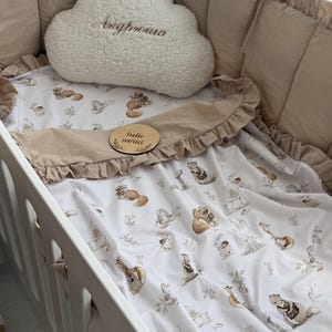Könnte beinhalten: Ein Babybett mit weißem Rahmen, mit einem wolkenförmigen Kissen und Bettwäsche mit einem Waldtiermuster. Die Bettwäsche hat einen Rüschenrand und eine Holzscheibe mit der Aufschrift "hello world".