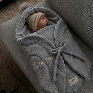 May include: A gray fleece baby swaddle blanket with a hood featuring a pom-pom. The swaddle has a tie closure, the embroidered name "Eloven" on the front, and a rectangular label with the text "PID ARYLEM" at the bottom right corner.