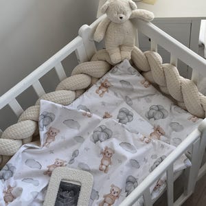 May include: A white crib with a cream-colored braided bumper and a patterned blanket featuring teddy bears and balloons. A plush teddy bear sits atop the crib, and a framed ultrasound image rests inside the crib.