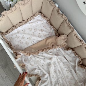 May include: A beige baby crib set with a patterned blanket and pillow featuring teddy bear illustrations. The set includes a padded bumper with ruffled edges. The crib is white.