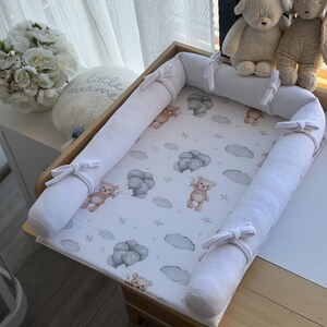 May include: A baby changing station with a light wood frame and white padded bumpers tied with bows. The changing pad features a pattern of teddy bears, clouds, and stars in soft gray tones. Two teddy bear plush toys sit on the edge.