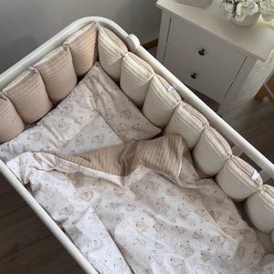 May include: A white crib featuring a set of padded bumpers in alternating beige and cream colors with a cable knit texture. The bedding set includes a white sheet and a blanket decorated with a repeating pattern of small bears and crescent moons.