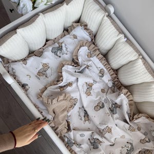 Könnte beinhalten: Ein weißes Babybett mit einer gemusterten Decke mit Cartoon-Tieren und Luftballons. Die Decke hat einen gerüschten Rand und eine beige Bordüre. Das Bettchen enthält auch weiße Strickkissen mit beigem Besatz.