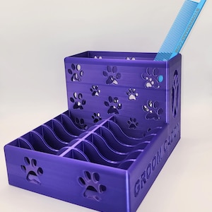 Op de afbeelding: Een paarse grooming caddy voor huisdieren met uitsparingen in de vorm van pootafdrukken en de tekst "GROOM CADDY" aan de zijkant. Een blauwe kam staat erin. De caddy heeft meerdere compartimenten.