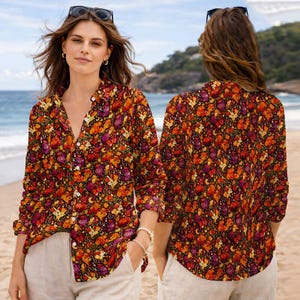 May include: A button-down shirt with a vibrant floral pattern in shades of red, orange, and purple. The long-sleeved shirt is paired with light-colored pants. Sunglasses are perched on the model's head. The background shows a beach and ocean.
