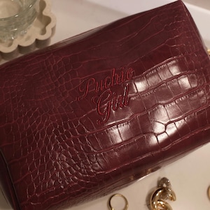 May include: A burgundy faux-leather clutch with a crocodile skin texture. The words "Puchie Girl" are embroidered in a matching burgundy thread. Gold jewelry, including a ring and a chain, is visible.