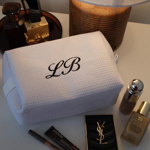 May include: White textured cosmetic bag with the embroidered initials "LB" in black. The bag is surrounded by makeup products, including a black compact with a gold logo, lip pencils, and foundation bottles. A lamp and perfume bottles are in the background.
