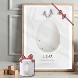 Könnte beinhalten: Ein gerahmter Druck und eine kleine dekorative Box mit einem Osterei-Design. Der Druck zeigt ein weißes Ei mit Hasenohren, den Namen "LINA" und den Text "Erstes Osterfest 2026". Ein rosa Band verziert den Rahmen und die Box.