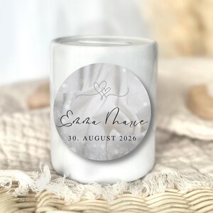 Könnte beinhalten: Eine weiße Keramikkerze mit einem runden Aufkleber. Der Aufkleber zeigt die Namen "Emma Marie" und das Datum "30. AUGUST 2026". Das Design umfasst zwei verschlungene Herzen und einen weichen, verschwommenen Hintergrund.
