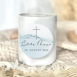 Könnte beinhalten: Eine weiße zylindrische Kerze mit einem runden Etikett. Das Etikett zeigt ein Kreuz, den Namen "Emma Marie" in Schreibschrift und das Datum "30. AUGUST 2026". Der Hintergrund des Etiketts hat ein blaues Wassermotiv.