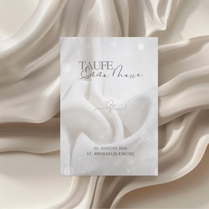 Könnte beinhalten: Eine weiße Einladungskarte mit dem Wort "TAUFE" oben, gefolgt von einem Namen in Schreibschrift und einem Herzdesign. Die Karte enthält auch das Datum "30. AUGUST 2026" und "ST. MICHAELIS KIRCHE" in schwarzem Text. Der Hintergrund ist ein weicher, drapierter Stoff.