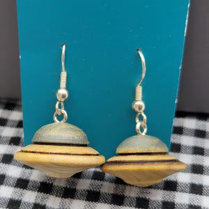 May include: A pair of wooden earrings with a silver hook. The earrings are shaped like flying saucers, with a light brown base and a gray top. The earrings are displayed against a teal background and a black and white checkered pattern.