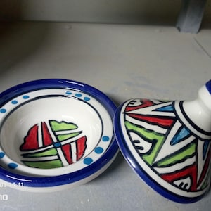Könnte beinhalten: Ein Keramik-Tajine-Gericht mit Deckel, mit weißer Basis und einem leuchtend blauen Rand. Das Gericht ist mit farbenfrohen geometrischen Mustern in Rot, Grün und Blau verziert. Der Deckel hat einen Griff und ist mit ähnlichen Mustern verziert.
