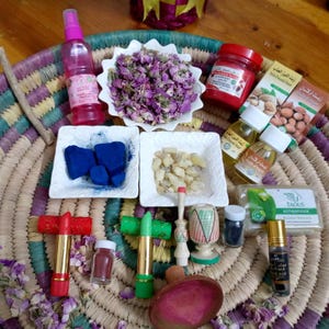 Puede incluir: Una colección de productos de belleza y bienestar, incluyendo pétalos de rosa secos, aceites esenciales, barras de labios e incienso, dispuestos sobre una cesta tejida. Los artículos están en varios recipientes y colores.