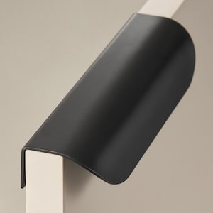 Op de afbeelding: Een zwarte kastgreep gemonteerd op een witte kastdeur. De handgreep heeft een gebogen ontwerp en is gemaakt van metaal. Het logo "Modern Brass Home Hardware" is zichtbaar aan de onderkant.