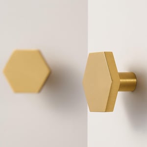 May include: Two gold-colored, hexagonal cabinet knobs. One knob is mounted on a white surface, while the other is floating in the background. The knobs have a modern design. The logo "Modern Brass Home Hardware" is at the bottom.