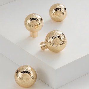 May include: Four gold-colored cabinet knobs with a hammered texture. The knobs are spherical and have a stem or a screw for attachment. The knobs are arranged on a white surface. The logo "Modern Brass Home Hardware" is visible.