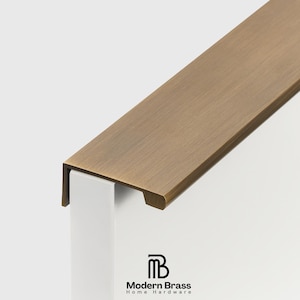 May include: A close-up of a bronze-colored metal door trim piece. The trim is rectangular with a slightly rounded edge and is mounted on a white support. The Modern Brass Home Hardware logo is visible.