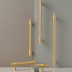 May include: A collection of gold-colored cabinet hardware, including handles and pulls, against a neutral gray and white backdrop. The hardware features a cylindrical design with decorative details. The Modern Brass Home Hardware logo is visible.