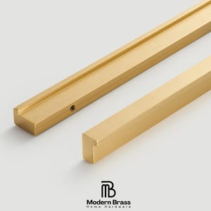 May include: Close-up of two gold-colored cabinet pulls. One pull is rectangular with a recessed channel, and the other is a solid, square bar. The Modern Brass logo is in the bottom right corner.