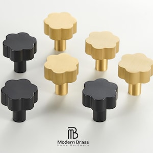 Może przedstawiać: Kolekcja gałek do szafek w kształcie kwiatów w kolorze czarnym i złotym. Gałki mają pięciopłatkowy kształt i cylindryczną podstawę. Obraz zawiera tekst "Modern Brass Home Hardware".