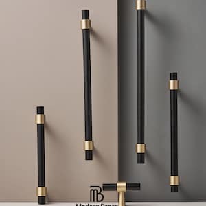 May include: A selection of black and gold cabinet pulls and handles. The pulls feature a cylindrical black body with gold-coloured accents. The handles are displayed against a neutral backdrop, highlighting their design. The Modern Brass Home Hardware logo is visible.