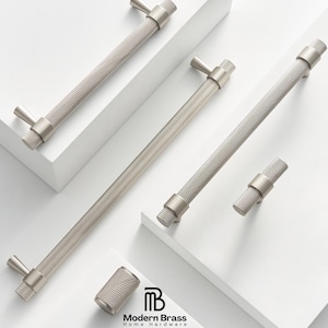 May include: A collection of silver-toned cabinet hardware, including handles and knobs, arranged on a white surface. The handles have a textured, diamond-patterned grip and a T-shaped end. The Modern Brass Home Hardware logo is visible.