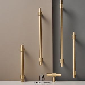 May include: A selection of gold-toned cabinet pulls and handles in different lengths. The hardware features a cylindrical design with a T-shaped end. The Modern Brass Home Hardware logo is at the bottom. The background is a neutral colour.