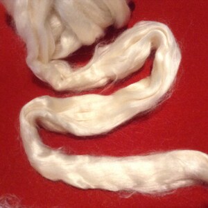 May include: A close-up of a white, fluffy, natural fiber, likely silk or cotton, arranged in a loose, flowing shape against a red background.