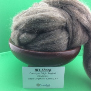 May include: A close-up of a light brown BFL sheep wool fiber in a wooden bowl. The wool is soft and fluffy, and the label says "BFL Sheep, Country of Origin: England, 26 Microns, Staple Length: 85-90mm (3.5")."