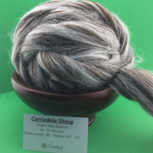 May include: A ball of Corriedale sheep wool in shades of gray, brown, and white, resting in a wooden bowl. The wool has a staple length of 3.5"-6" and originates from New Zealand. A tag with details is visible.