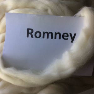 May include: A close-up of a bundle of off-white Romney wool roving. A white card with the word "Romney" in black lettering is partially visible within the wool. The wool appears soft and fluffy, ideal for fiber arts.