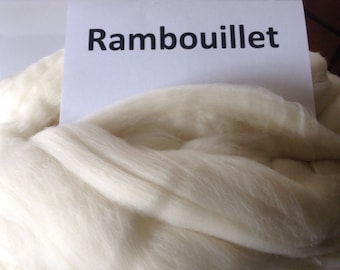 May include: Close-up of a pile of off-white Rambouillet wool fiber. The fiber is soft and fluffy, with a slightly crimped texture. A white card with the word "Rambouillet" in black text is placed on top of the wool.