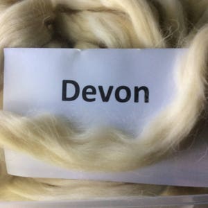 May include: Close-up of a pile of light beige wool fibers. A white label with the word "Devon" in black text is placed on top of the wool. The wool fibers are soft and fluffy, suggesting a textile product.