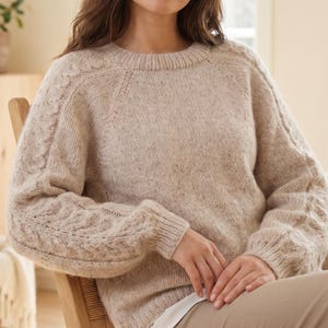 May include: A beige, speckled, long-sleeved knit jumper featuring a crew neckline and cable-knit detailing along the raglan sleeves. The garment is crafted from a textured, fuzzy yarn and includes ribbed cuffs and a ribbed hem for a structured fit.