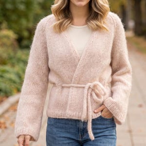 Könnte beinhalten: Eine hellrosa, langärmelige Wickelstrickjacke aus flauschigem Strickgarn. Die Strickjacke verfügt über einen V-Ausschnitt und einen passenden Bindegürtel in der Taille, kombiniert mit einer blauen Jeans und einem hellen Unterziehshirt.
